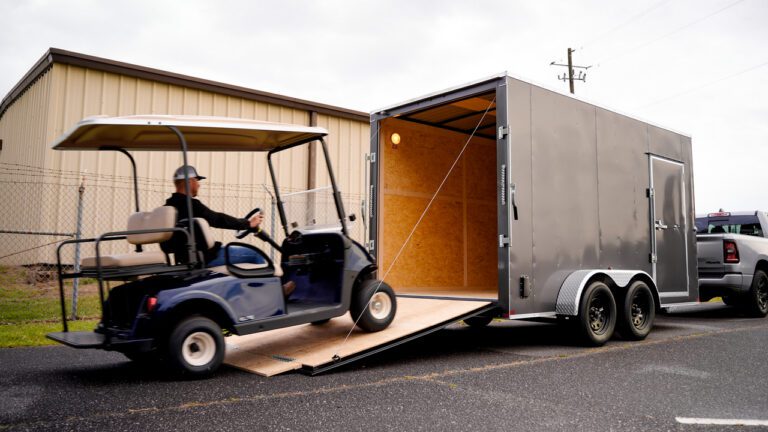 Golf cart driving up a ramp into an open enclosed trailer attached to a pickup truck on a roadside.