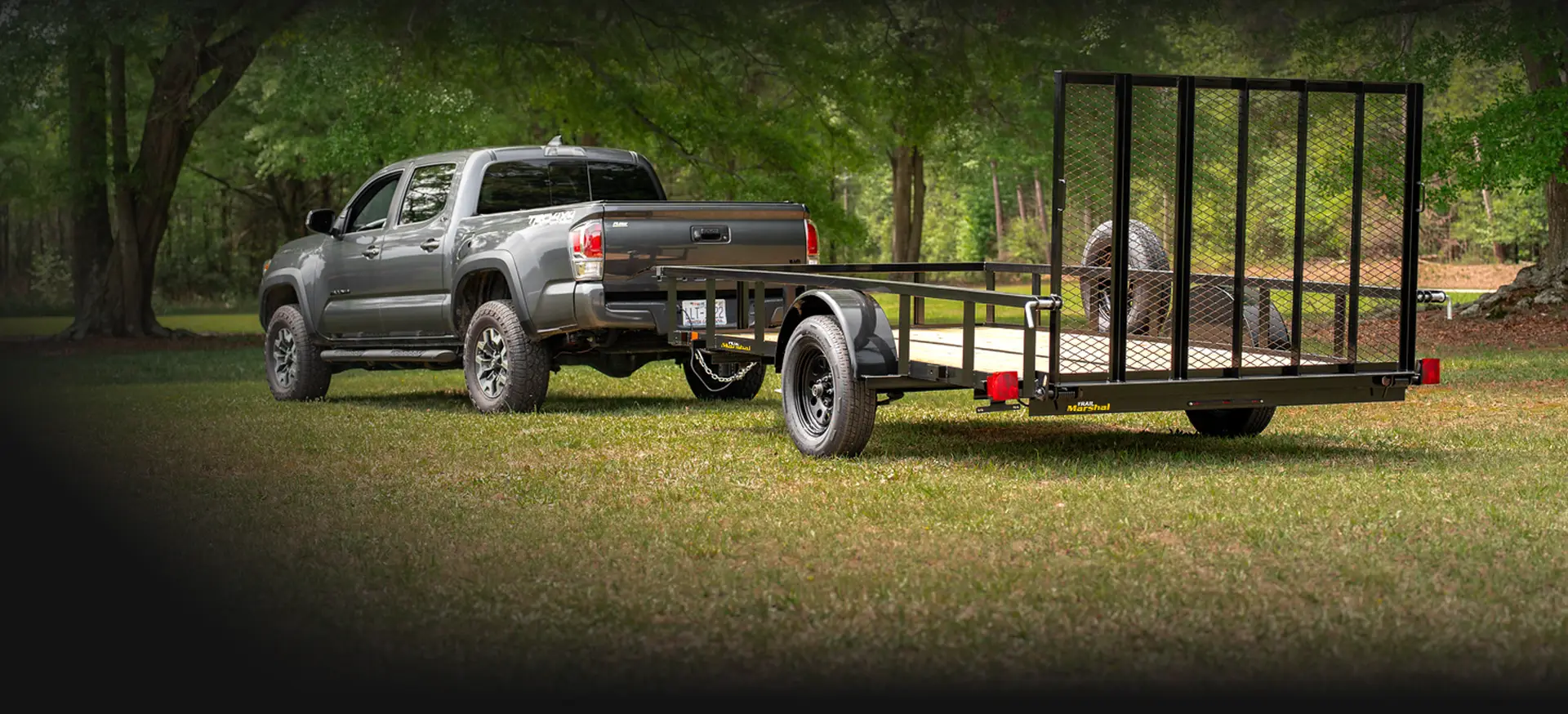 Gray pickup truck towing a black single-axle utility trailer with mesh rear ramp gate, parked on a grassy field beneath large trees. A spare tire is mounted at the front of the trailer, and the open trailer bed is empty.