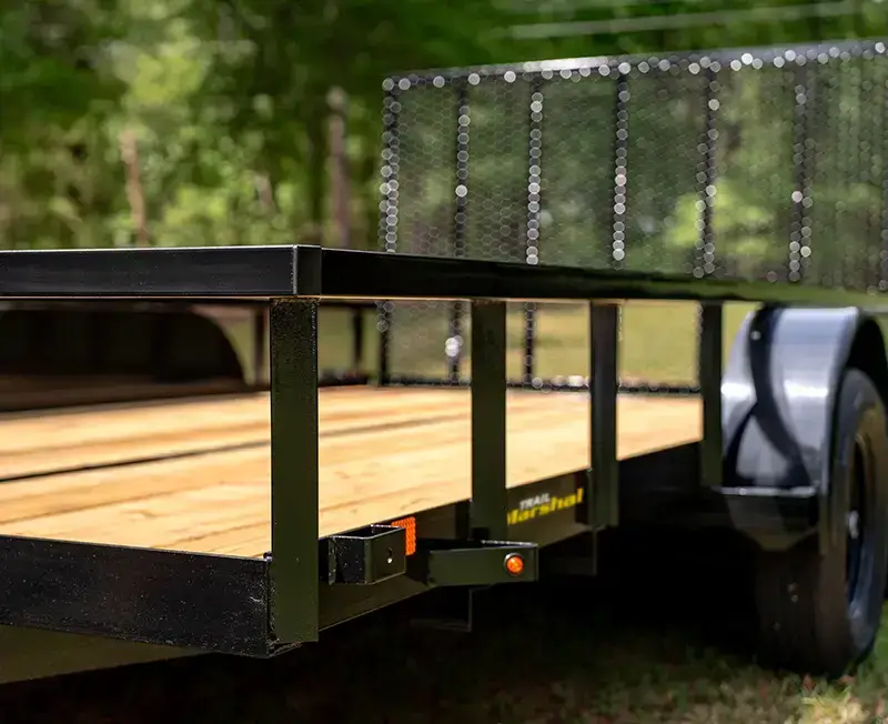 Close-up of a black utility trailer with a wood plank deck, steel side rails, mesh front panel, and single axle, shown outdoors on grass.