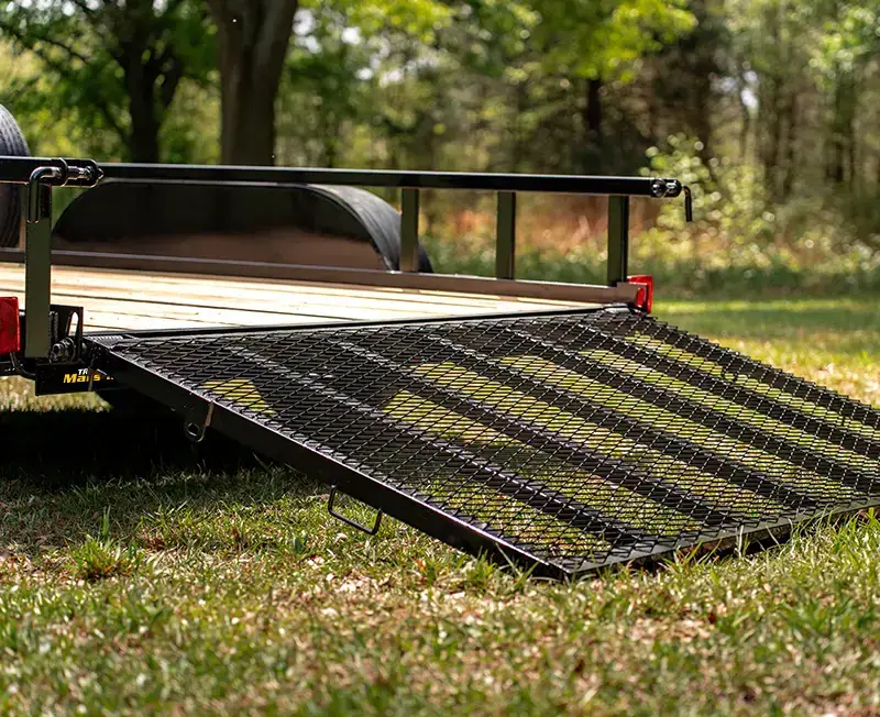 Rear view of a utility trailer with the expanded metal loading ramp lowered onto the grass, showing the wood deck, side rails, and rear lights.
